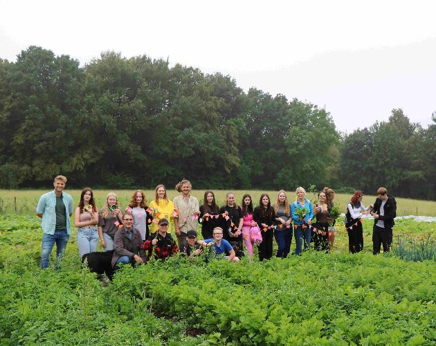 Auf dem Bild ist eine aufgereihte Schülergruppe mit Lehrer*innen und Mitarbeitenden der Solawi Mülheim inmitten eines Feldes zu sehen. Die fotografierten Personen halten eine Girlande und verschiedene Pflanzen in den Händen. 
